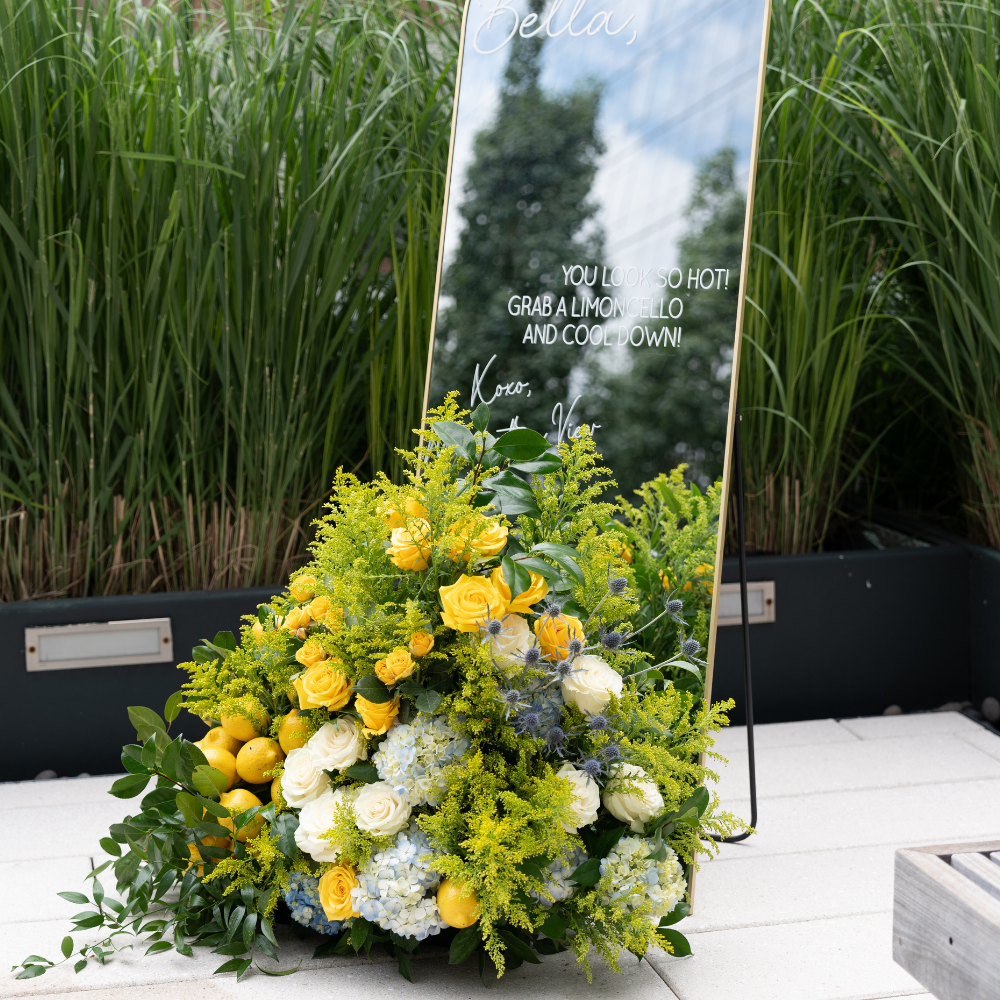 Vibrant low centerpiece floral arrangement reflecting limoncello theme in yellow, green, and blue featuring hydrangea, roses, blue thistle, lemons, and greenery, displayed in front of an event mirror